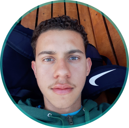 Foto do Pablo Perdigão com bigode,usando uma jaqueta verde escura,com um fundo de madeira ripada e uma mochila da nike