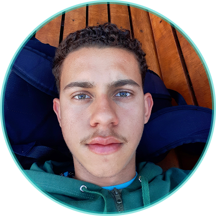 Foto do Pablo Perdigão com bigode,usando uma jaqueta verde escura,com um fundo de madeira ripada e uma mochila da nike